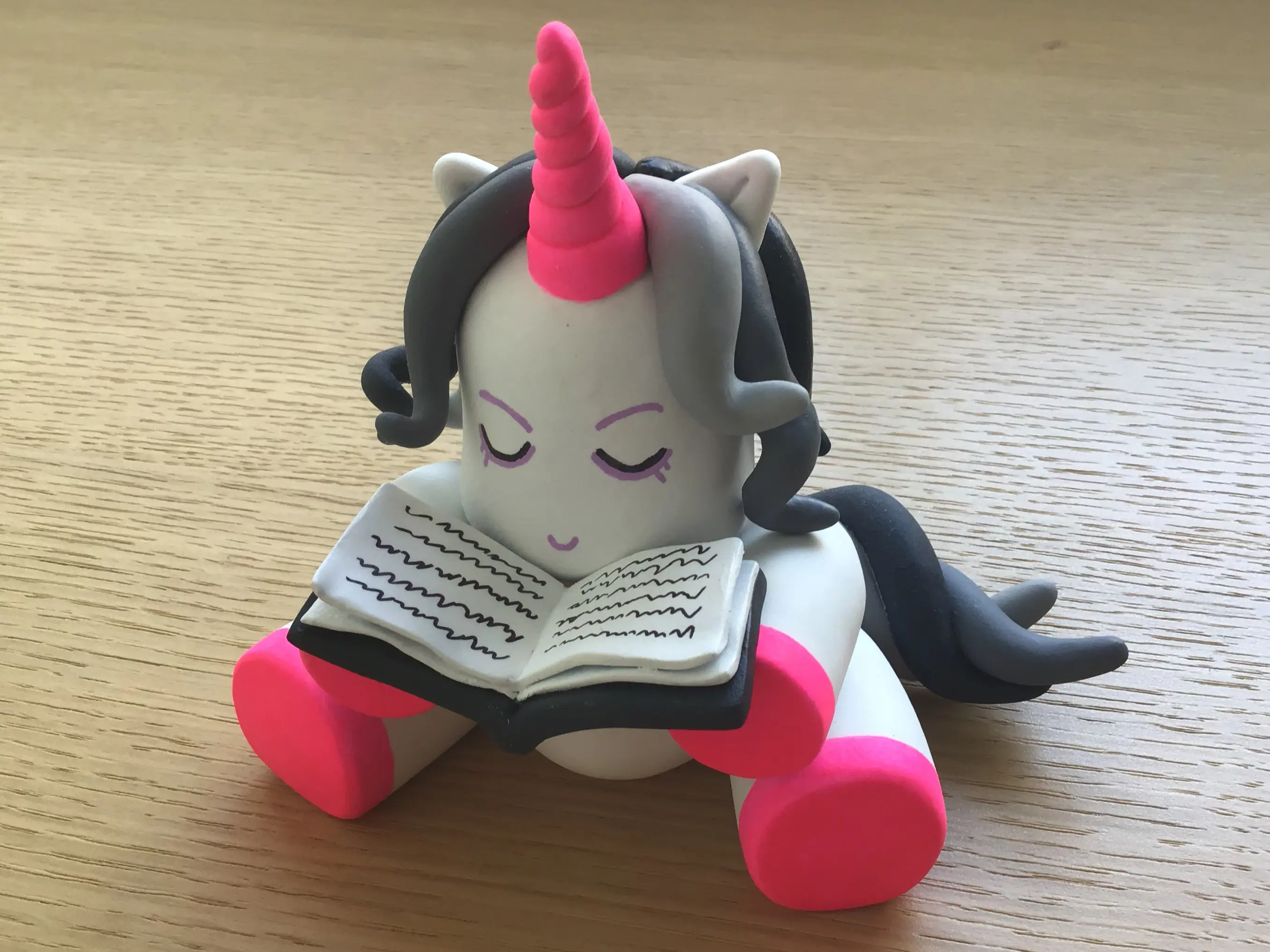 Figura de arcilla polimérica, Art Toy, terminada. Representa un unicornio sentado con un libro abierto en sus manos.